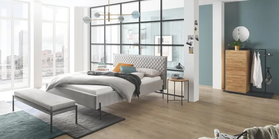 Schlafzimmer mit grauem Polsterbett, Sitzbank und Holzkommode vor einer Glaswand.